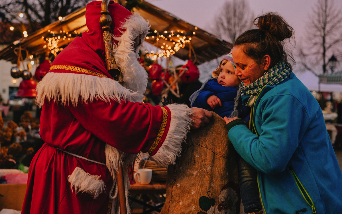 Weihnachtsmann gibt Geschenk an Mutter mit Kind auf Weihnachtsmarkt.