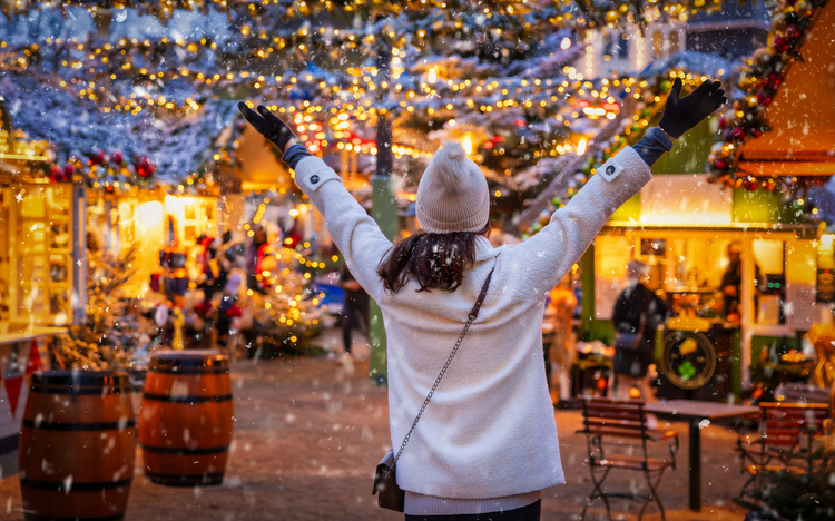 Person auf Weihnachtsmarkt, Hände hoch, umgeben von Lichtern und Dekorationen.