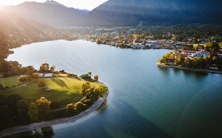 Luftaufnahme eines Sees mit umliegender Landschaft und Bergen im Hintergrund bei Sonnenuntergang