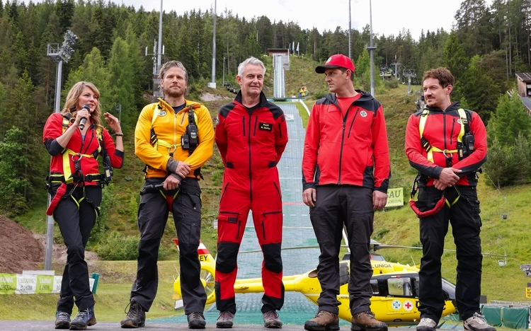 Fünf Personen in Outdoor-Kleidung vor grüner Waldkulisse und Skisprungschanze.