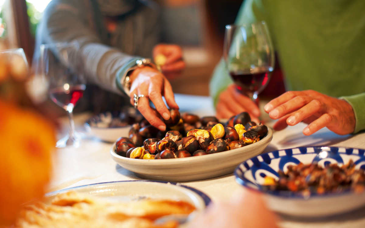 Personen greifen nach Maronen auf einem gedeckten Tisch mit Wein und Tellern.