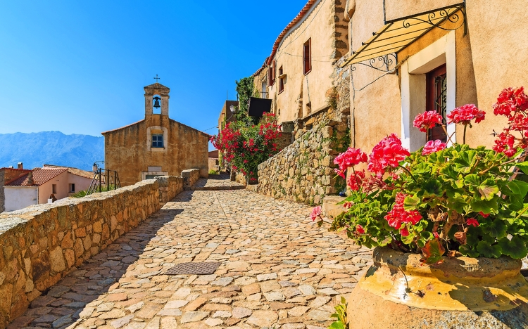 Steinpflasterstraße in mediterranem Dorf mit Kirche und blühenden Geranien.